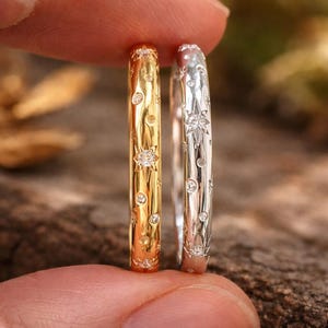 May include: Two rings, one gold and one silver, are held between fingers. Both rings feature a repeating pattern of star and dot designs. The gold ring has a warm, shiny finish, while the silver ring has a bright, polished appearance. The rings are likely jewelry.