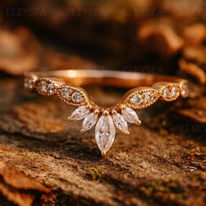 Puede incluir: Un anillo de oro rosa con un diseño curvo, con una fila de pequeñas piedras claras redondas y un grupo central de piedras con corte marquesa. El anillo se encuentra sobre un fondo natural borroso.