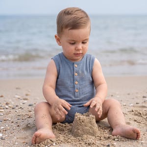 Può includere: Una tutina senza maniche blu in tessuto di mussola testurizzato con scollo rotondo e un'abbottonatura frontale con quattro piccoli bottoni in legno. Il capo è indossato da un bambino seduto su una spiaggia sabbiosa mentre gioca con un castello.