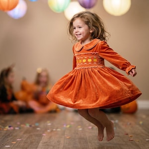 May include: A long-sleeved orange velvet dress featuring a Peter Pan collar and a smocked bodice with embroidered yellow and orange pumpkin motifs. The dress has a flared skirt and ruffled detailing along the chest and sleeve cuffs.