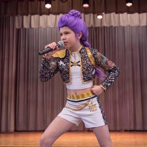 May include: A performer wearing a white crop top and matching shorts with gold trim, paired with a black sequined jacket featuring gold accents. The outfit includes a gold chain belt detail. The performer has styled purple hair and holds a microphone.