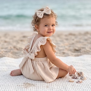 May include: A beige muslin romper featuring ruffled shoulder straps and a tie-back closure, paired with a matching beige bow headband. The garment is crafted from a textured, lightweight fabric with a soft, neutral tone and a simple, open-back design.