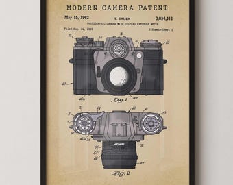 May include: Framed print of a vintage camera patent. The print features two detailed diagrams of a camera, labeled "Fig. 1" and "Fig. 2", with technical annotations. The text reads "MODERN CAMERA PATENT" and "May 15, 1962".