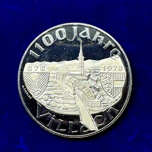 Könnte beinhalten: Eine silberne Gedenkmünze mit dem Text "1100 Jahre" und den Jahreszahlen "878" und "1978". Die Münze zeigt eine detaillierte Darstellung einer Stadt mit Kirche und Brücke. Die Münze ist vor einem dunkelblauen Hintergrund platziert.