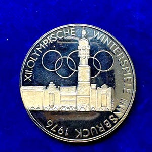 May include: A silver coin with the text "XII OLYMPISCHE WINTERSPIELE IN INNSBRUCK 1976" encircling the edge. The coin features the Olympic rings and a building design. The background is a deep blue.