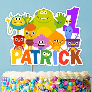 Puede incluir: Un adorno para pastel colorido con un grupo de monstruos de dibujos animados y el nombre "PATRICK" en letras grandes y multicolores. También se incluye un gran número "1" púrpura, lo que sugiere un tema de primer cumpleaños. El pastel está decorado con glaseado blanco y chispas coloridas.