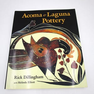 May include: A book titled "Acoma & Laguna Pottery" by Rick Dillingham with Melinda Elliott. The cover features a black background with a stylized illustration in brown, orange, and black, with floral and bird-like motifs.
