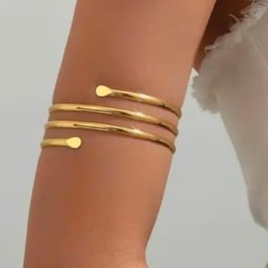 May include: A gold-toned arm cuff featuring a triple-coil design that wraps around the upper arm. The metal band has a smooth, polished finish with rounded, tapered ends at each opening, creating a continuous spiral appearance on the skin.