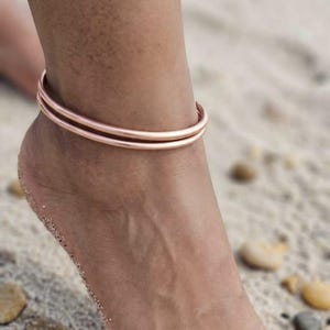 Puede incluir: Una tobillera de doble banda en oro rosa en un tobillo. La tobillera se lleva en un pie en una playa arenosa con pequeños guijarros. La tobillera tiene un diseño simple y elegante.