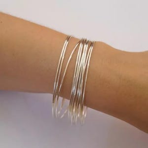 May include: A collection of thin, silver bangles stacked on a wrist. The bangles are simple and elegant, reflecting light. The jewellery is a minimalist accessory.