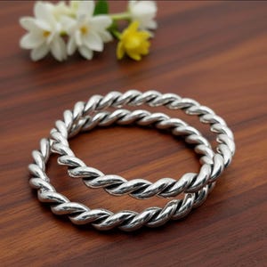 May include: Two silver-colored, twisted rope design bangles. The bangles are circular and sit on a dark brown wooden surface. White and yellow flowers are in the background.