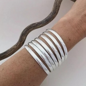 Op de afbeelding: Een stapel glanzende, zilverkleurige armbanden om een pols. De armbanden zijn glad en afgerond en reflecteren licht. De sieraden hebben een eenvoudig, elegant ontwerp. Een gebogen, bruine tak staat op de achtergrond.
