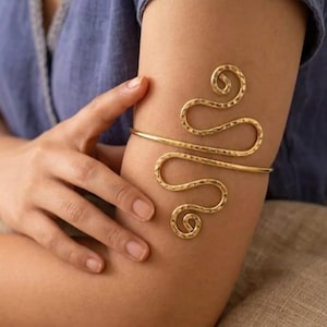 May include: A gold-toned metal arm cuff featuring a hammered texture and a continuous, winding wire design that forms two symmetrical, swirling loops extending from a central horizontal band that wraps around the upper arm.