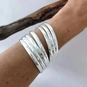 May include: A stack of silver bangles with a hammered texture. The bracelets are worn on a person's arm. The background is a light color with a wooden branch.