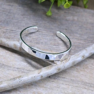 May include: A silver cuff bracelet with a faceted design. The open-ended bracelet is displayed on a light-colored wooden surface with a small branch and green leaves in the background. The bracelet is approximately 0.25 inches wide.