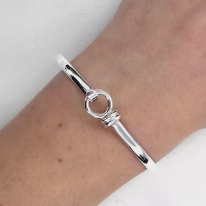 Peut inclure: Un bracelet jonc argenté brillant avec un fermoir circulaire. Le bracelet est lisse et poli, reflétant la lumière. Le fermoir présente un petit anneau et des détails décoratifs. Le bracelet est porté au poignet.