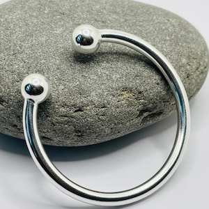 May include: A polished silver cuff bracelet with a minimalist design. The bracelet features a smooth, curved band that terminates in two spherical, ball-shaped ends. The bracelet is shown on a gray stone surface.