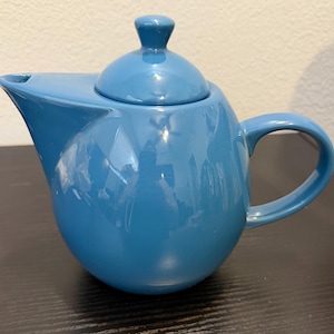 Könnte beinhalten: Eine glänzende, mittelblaue Keramik-Teekanne mit rundem Körper, einem geschwungenen Griff und einem passenden Deckel mit kleinem Knauf. Die Teekanne verfügt über einen breiten, integrierten Ausguss und eine glatte, reflektierende Oberfläche.