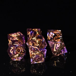 Puede incluir: Un conjunto de siete dados poliédricos en morado y dorado. Los dados son transparentes, con números dorados y elementos decorativos internos. Estos dados están diseñados para juegos de rol de mesa.