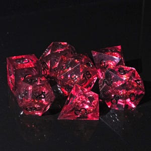 Op de afbeelding: Set van zeven rode en zwarte polyhedrale dobbelstenen voor tabletopspellen. De dobbelstenen zijn transparant met een glitterinterieur en zwarte nummering. Inclusief een D4, D6, D8, D10, D12, D20 en een percentagedobbelsteen.