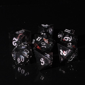 May include: A set of seven polyhedral dice, each with a clear, black, and red design. The dice have white numbering and are designed for tabletop role-playing games. The dice are displayed on a reflective black surface.