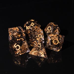 May include: Set of seven polyhedral dice with a black resin base and gold leaf inclusions. Each die features gold numbering. The dice are arranged on a reflective black surface.