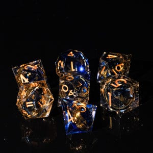Puede incluir: Un conjunto de siete dados poliédricos transparentes con interior azul y dorado y numeración dorada. Los dados están dispuestos sobre una superficie negra, con la luz reflejada en los dados. Ideales para juegos de mesa.