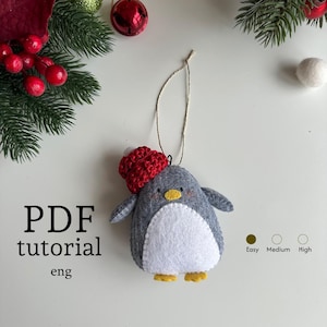 Puede incluir: Adorno navideño de pingüino de fieltro hecho a mano con gorro rojo, cuerpo gris y vientre blanco. El pingüino tiene patas amarillas y pico amarillo. El texto "PDF tutorial eng" es visible. El nivel de dificultad se indica como fácil.