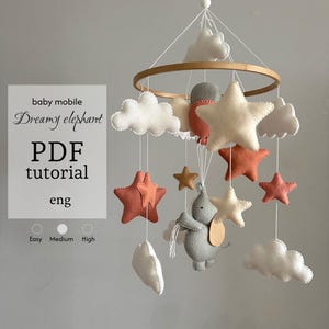 Könnte beinhalten: Ein Baby-Mobile PDF-Tutorial mit dem Titel Dreamy elephant, bestehend aus einem runden Holzrahmen mit hängenden Filzfiguren wie einem grauen Elefanten, weißen Wolken und Sternen in Creme und Terrakotta an weißen Schnüren.