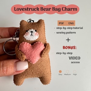 May include: A brown felt bear bag charm holding a pink heart. The bear has a white face and black eyes. The charm includes a metal keyring. Text on the image reads "Lovestruck Bear Bag Charm" and details about a PDF tutorial and video access.