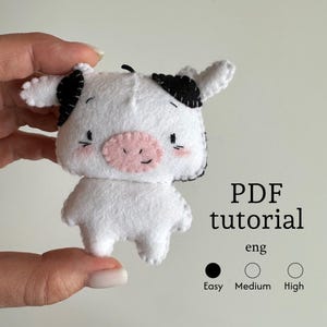 May include: A handmade white felt cow with black spots on its ears and head, and a pink nose. The cow has a smiling face with small black eyes and blush. The image includes the text "PDF tutorial" and difficulty levels.