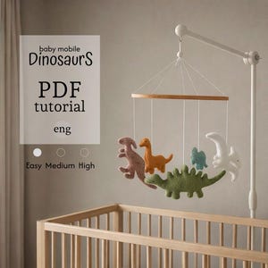 Può includere: Un mobile per bambini con dinosauri in feltro rosa, arancione, verde, blu e bianco, sospesi a un anello di legno. Il mobile è appeso sopra una culla di legno. Il testo a sinistra recita "baby mobile Dinosaurs PDF tutorial eng".