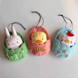 May include: Three Easter-themed felt ornaments. A white bunny in a green egg with carrot embroidery, a yellow chick in a pink egg with flower embroidery, and a white chick with a red heart in a blue egg with flower embroidery. Each has a black string.