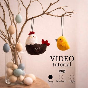 May include: An Easter-themed display featuring felt ornaments and eggs. A brown and white chicken and a yellow chick hang from a branch. The display includes a glass vase with eggs and the text "VIDEO tutorial eng".