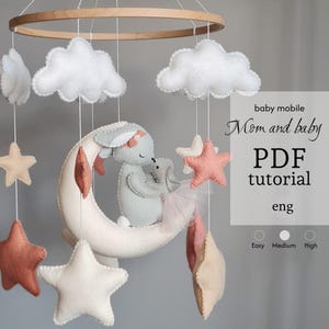 Puede incluir: Móvil para bebé con un conejito de fieltro y un conejito más pequeño abrazándose en una luna creciente. El móvil incluye nubes blancas, estrellas de varios colores y el texto "baby mobile Mom and baby PDF tutorial eng".