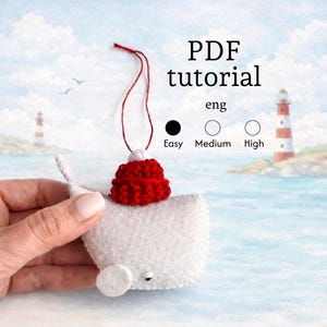 Puede incluir: Adorno de ballena de fieltro blanco con un gorro rojo tejido y una cuerda roja para colgar. El adorno tiene un ojo pequeño y una aleta redonda. La imagen incluye el texto "PDF tutorial" y niveles de dificultad: Fácil, Medio y Alto.