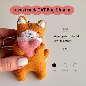 Könnte beinhalten: Orangener Filz-Katzen-Taschenanhänger, der ein rosa Herz hält. Die Katze hat weiße Akzente an Ohren und Gesicht, mit schwarzen Nahtdetails. Der Text auf dem Bild lautet "Lovestruck CAT Bag Charm", "PDF" und "ENG".