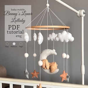 May include: A baby mobile featuring a wooden ring with hanging felt shapes. The mobile includes white clouds, felt stars, and a crescent moon with a sleeping bunny. The text "Bunny's Lunar Lullaby" is visible.