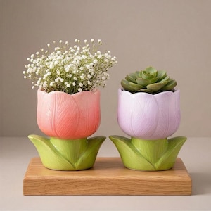 Peut inclure: Deux pots en céramique en forme de tulipe, un rose et un lavande, chacun avec des bases en forme de feuilles vertes. Le pot rose contient des fleurs blanches, et le pot lavande contient une plante succulente verte. Les pots sont sur un plateau en bois.