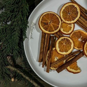 Könnte beinhalten: Ein weißer Teller enthält getrocknete Orangenscheiben und Zimtstangen. Die Orangenscheiben sind kreisförmig und hellbraun. Die Zimtstangen sind lang und braun. Grüne Kiefernzweige befinden sich im Hintergrund.