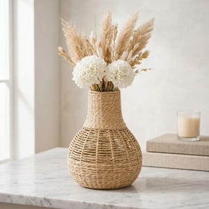 May include: A beige woven vase with a rounded base and a narrow neck, containing dried beige pampas grass and white flowers. A lit candle in a glass and two books are to the right of the vase.