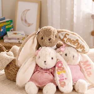 Puede incluir: Tres conejos de peluche con orejas largas. El conejo superior es marrón. El conejo inferior izquierdo es blanco con un vestido de cuadros rosas. El conejo inferior derecho es blanco con una blusa rosa. Las orejas tienen bordados los nombres Evelyn, Olivia y Mia.