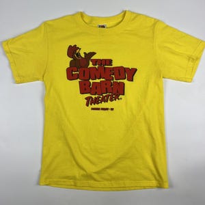 May include: Yellow t-shirt featuring the text "THE COMEDY BARN THEATER" in red and brown. The shirt has a crew neck and short sleeves. The graphic includes a cartoon character. The shirt is made of a soft material.