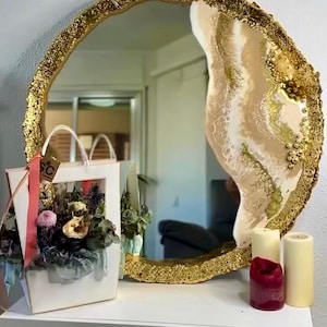 Op de afbeelding: Een ronde decoratieve spiegel met een goudkleurige, getextureerde lijst. De binnenkant van de spiegel heeft een wit en beige ontwerp. Een witte cadeauzak met bloemen en kaarsen staat op een wit oppervlak voor de spiegel.