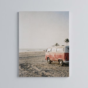 Op de afbeelding: Een rode en witte vintage Volkswagenbus geparkeerd op een zandstrand. De bus staat met het gezicht naar de oceaan, met palmbomen op de achtergrond. De afbeelding heeft een warme, nostalgische uitstraling, die reizen en avontuur oproept.