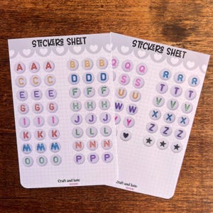 Peut inclure: Deux feuilles d'autocollants avec des lettres de l'alphabet colorées. Chaque lettre est dans un cercle. Les feuilles sont étiquetées "STICKERS SHEET" en haut et "Craft and latte CLUB" en bas.