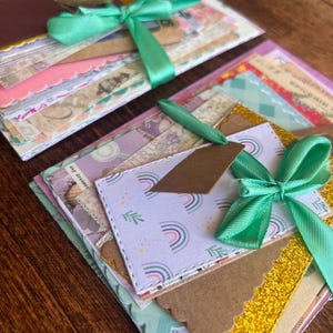 Peut inclure: Deux piles d'éphémères en papier faits à la main, nouées avec un ruban de satin vert menthe. Les piles comprennent divers papiers à motifs, étiquettes et cartes avec des bords festonnés, des arcs-en-ciel et des accents de paillettes dorées.