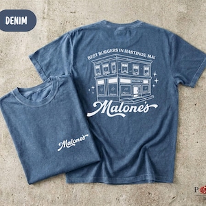 May include: A denim blue short-sleeve t-shirt featuring a white graphic on the back of a building with the text "BEST BURGERS IN HASTINGS, MA!" and "Malone's". The front chest area displays the white script text "Malone's" on the left side.