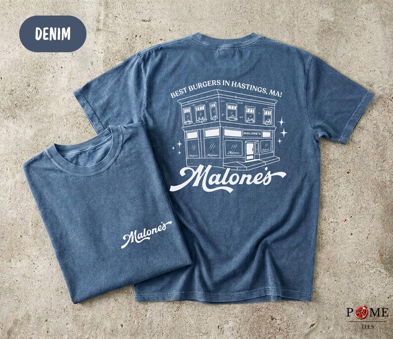 May include: A denim blue short-sleeve t-shirt featuring a white graphic on the back of a building with the text "BEST BURGERS IN HASTINGS, MA!" and "Malone's". The front chest area displays the white script text "Malone's" on the left side.