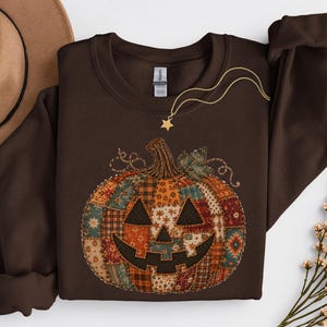May include: A dark brown crewneck sweatshirt featuring a large, central graphic of a jack-o'-lantern pumpkin. The pumpkin is designed with a patchwork pattern of various autumn-toned fabrics, including plaid, floral, and geometric prints in orange, red, and teal.